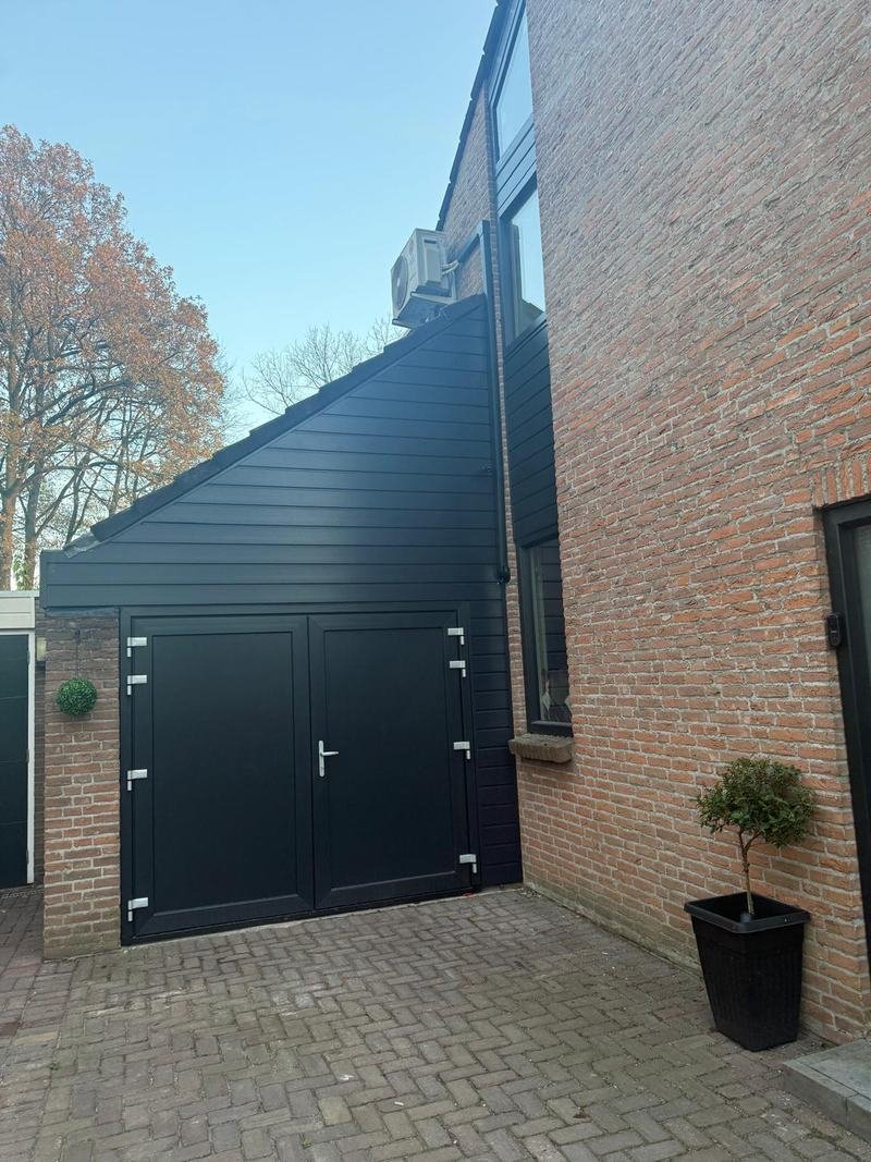 Garage met nieuwe zwarte gevelbekleding en dubbele deuren naast een bakstenen woning
